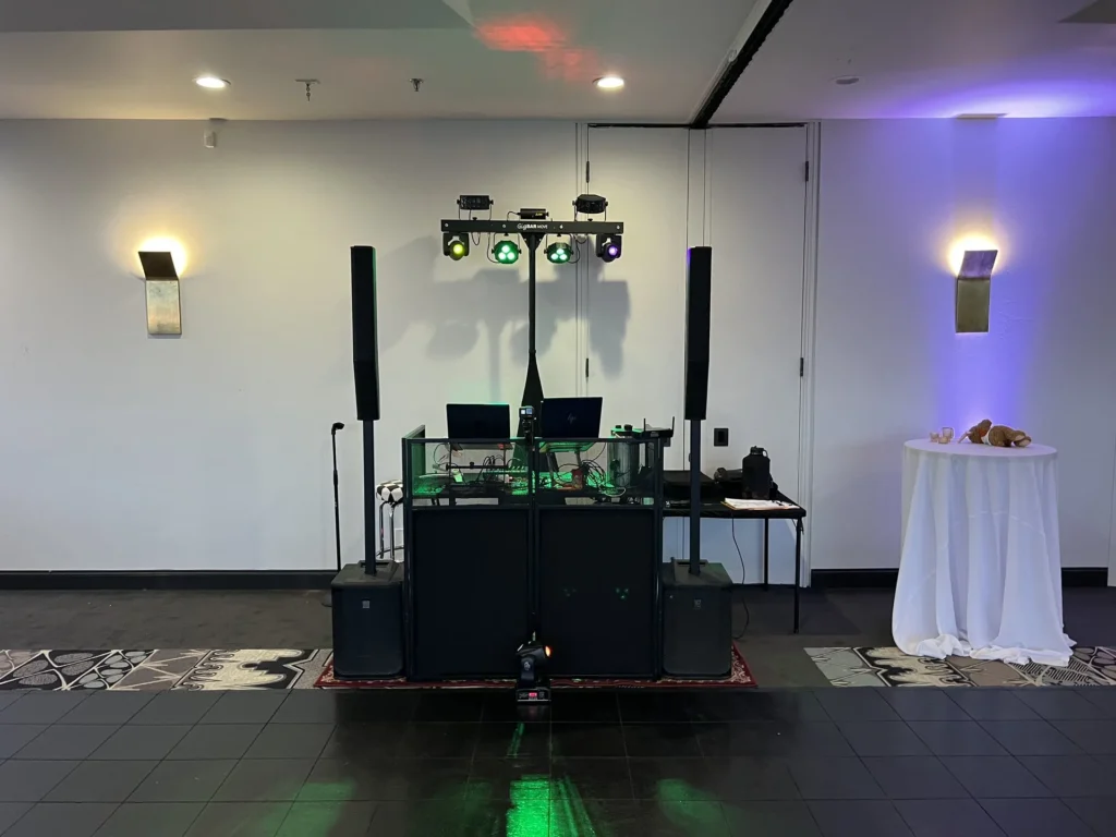 DJ booth, speaker stacks and lighting trees set up in an empty ballroom with purple uplighting