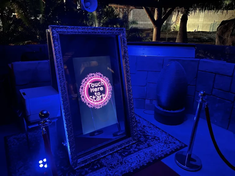 Mirror photo booth glowing blue on an outdoor patio at night with a Touch Here To Start screen