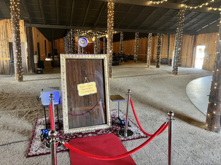 Mirror photo booth with a red carpet and stanchions set up inside a barn venue strung with lights