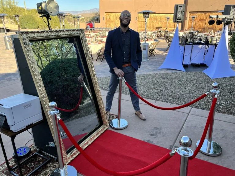 Mirror photo booth with red carpet and rope stanchions set up outdoors beside the attendant